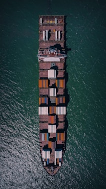 An aerial view of a large cargo ship fully loaded with colorful containers, navigating through blue-green ocean waters. The vessel's rectangular shape and container arrangement are clearly visible from above.