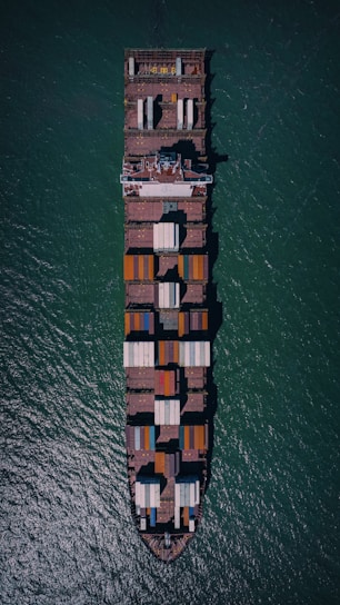 An aerial view of a large cargo ship fully loaded with colorful containers, navigating through blue-green ocean waters. The vessel's rectangular shape and container arrangement are clearly visible from above.