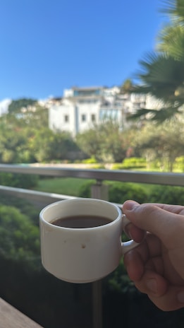 A hand reaching for a freshly brewed cup of coffee, with a backdrop of lush greenery visible through a window.