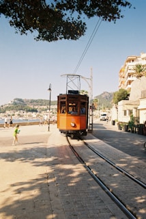 A vintage orange tram travels along a cobblestone street lined with tracks. Shadows of leaves cover part of the pavement while a child in green shorts walks nearby. The background features a coastal town with people strolling along the promenade. Hills and a clear blue sky can be seen in the distance, with buildings on the right.