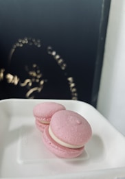 Two pink macarons rest on a white paper tray. The delicate cookies have a smooth texture with creamy filling visible between the layers. A dark, blurry background with faint text and design elements is visible in the upper part of the image.