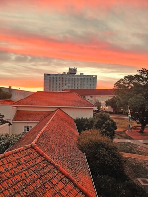 A dramatic sunset paints the sky with vibrant hues of orange, red, and pink above a cityscape. The foreground features rooftops with reddish-orange tiles and a few buildings with different architectural styles. A tall building stands prominently in the background, while surrounding trees and pathways can be seen. The South African flag is visible, and two people are walking near it.