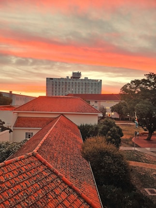 A dramatic sunset paints the sky with vibrant hues of orange, red, and pink above a cityscape. The foreground features rooftops with reddish-orange tiles and a few buildings with different architectural styles. A tall building stands prominently in the background, while surrounding trees and pathways can be seen. The South African flag is visible, and two people are walking near it.