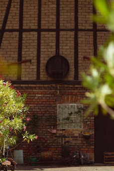 A rustic brick wall with wooden beams and a round wooden barrel attached at the top. Below, a variety of gardening tools, colorful watering cans, and vintage items are arranged against the wall. Bright green foliage with pink flowers partially frames the scene.