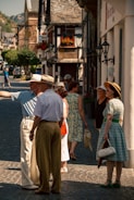 A picturesque street scene in a quaint village with people wearing vintage-style clothing. The cobblestone path is lined with charming, old European buildings adorned with flowers. A group of people is gathered and appears to be enjoying a leisurely conversation.