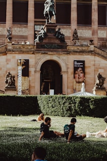 A grand museum building with classical architecture, including columns and statues, as well as banners displaying artwork. In the foreground, people are relaxing on a grassy area surrounded by well-manicured hedges.