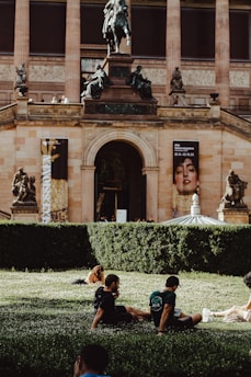 A grand museum building with classical architecture, including columns and statues, as well as banners displaying artwork. In the foreground, people are relaxing on a grassy area surrounded by well-manicured hedges.