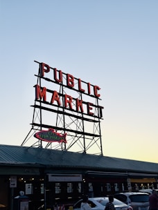 Large metal sign suspended above a building with the words 'PUBLIC MARKET' in bright red letters. Below is a smaller sign shaped like a fish with the words 'CITY FISH MARKET'. The market appears to be on a street with parked cars and a few people walking by. The sky is clear, indicating it's either early morning or late afternoon.