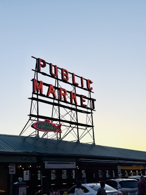 Large metal sign suspended above a building with the words 'PUBLIC MARKET' in bright red letters. Below is a smaller sign shaped like a fish with the words 'CITY FISH MARKET'. The market appears to be on a street with parked cars and a few people walking by. The sky is clear, indicating it's either early morning or late afternoon.
