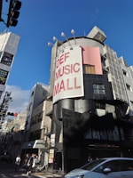 A street scene featuring a tall building with a large sign reading 'DEF MUSIC MALL'. The building has a modern design with a mix of black and gray exterior colors, along with a pink section. There are people walking on the sidewalk and a parked bicycle near the entrance of the building. Several streetlights and other buildings are visible around the area. The sky is clear and blue, suggesting a sunny day.