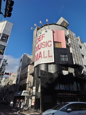 A street scene featuring a tall building with a large sign reading 'DEF MUSIC MALL'. The building has a modern design with a mix of black and gray exterior colors, along with a pink section. There are people walking on the sidewalk and a parked bicycle near the entrance of the building. Several streetlights and other buildings are visible around the area. The sky is clear and blue, suggesting a sunny day.