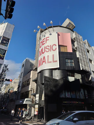 A street scene featuring a tall building with a large sign reading 'DEF MUSIC MALL'. The building has a modern design with a mix of black and gray exterior colors, along with a pink section. There are people walking on the sidewalk and a parked bicycle near the entrance of the building. Several streetlights and other buildings are visible around the area. The sky is clear and blue, suggesting a sunny day.