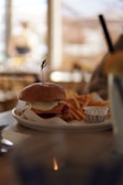 A plate with a burger topped with cheese, accompanied by a serving of fries and a small cup of sauce. The scene is set on a wooden table, with soft, blurred background suggesting a cozy cafe atmosphere.