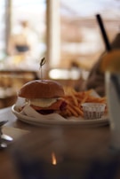 A plate with a burger topped with cheese, accompanied by a serving of fries and a small cup of sauce. The scene is set on a wooden table, with soft, blurred background suggesting a cozy cafe atmosphere.
