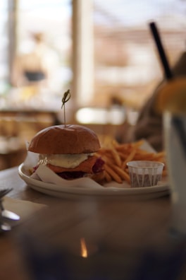 A plate with a burger topped with cheese, accompanied by a serving of fries and a small cup of sauce. The scene is set on a wooden table, with soft, blurred background suggesting a cozy cafe atmosphere.