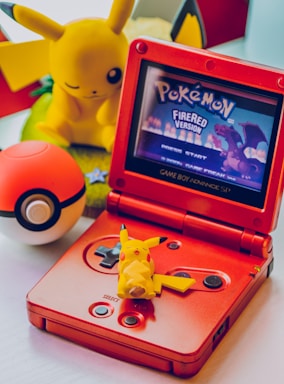 A red Game Boy Advance SP displays the title screen of 'Pokemon FireRed Version.' A small Pikachu figure is placed on the game console. In the background, a larger Pikachu plush toy and a Pokeball are visible.