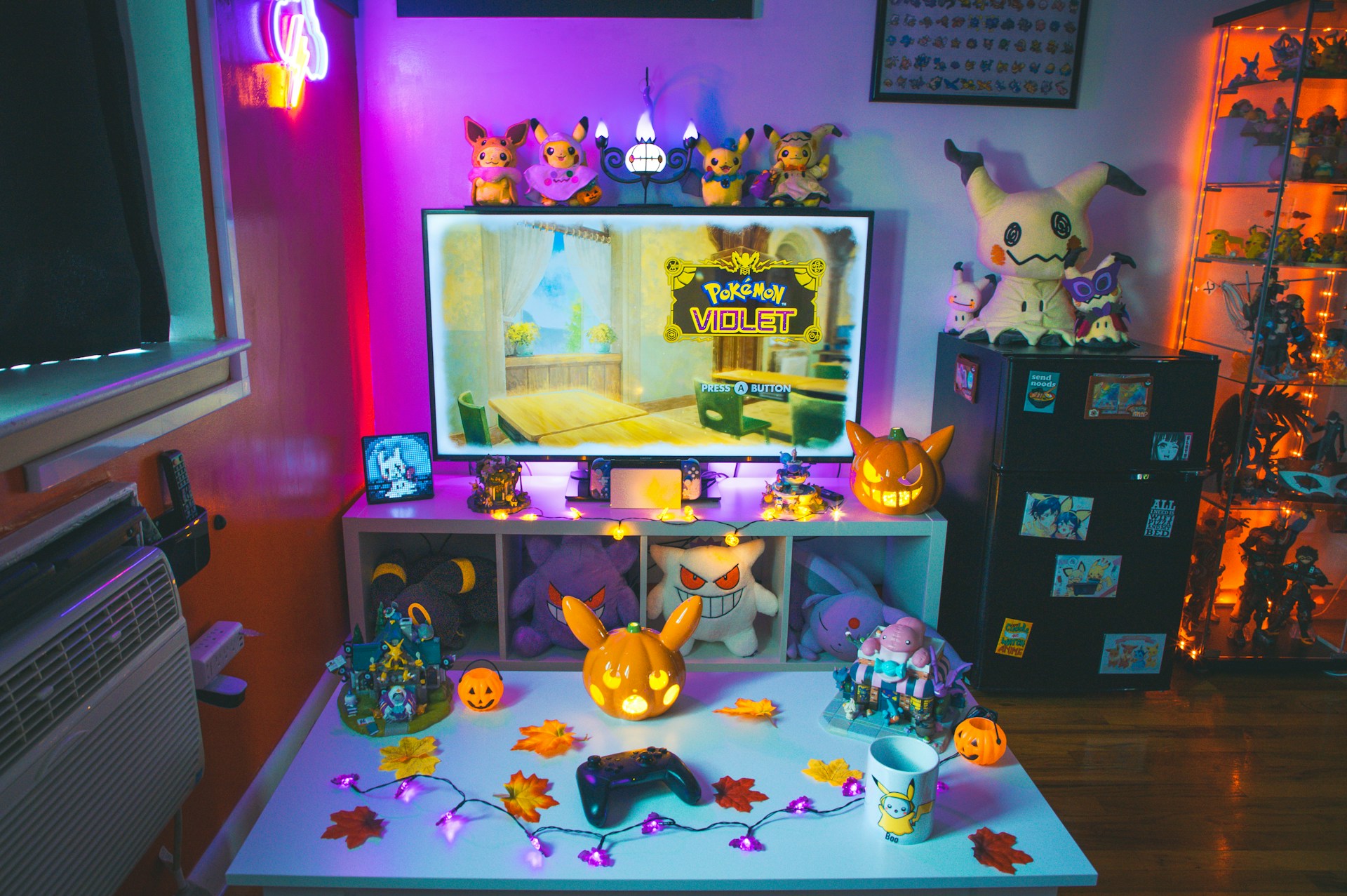 The room is decorated with various Pokemon-themed items, including plush toys, figurines, and a lit-up jack-o'-lantern. The center features a TV displaying the title screen of Pokemon Violet, with colorful lighting and autumn-themed decorations, such as artificial leaves and string lights, in the foreground. Shelving units hold more Pokemon collectibles.