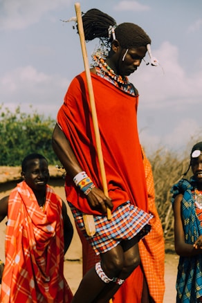 A group of people dressed in traditional attire featuring vibrant reds, blues, and patterned fabrics. One individual, likely engaged in a cultural dance or ceremony, is holding a long stick and is adorned with intricate beadwork and hair decorations. The background showcases a natural setting with foliage under a partly cloudy sky.
