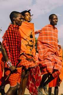 Three people are dressed in colorful red and orange traditional attire, participating in what appears to be a cultural or ceremonial event. They are energetically jumping, with vibrant patterns and beaded accessories visible. The background shows a clear sky.