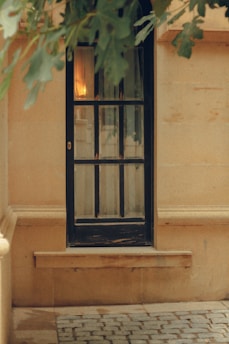 A wooden framed glass door with multiple panes set against a stone wall. Above the door, leaves from a nearby tree gently hang. Soft light from inside illuminates the glass, creating a warm glow. The ground is paved with cobblestones.