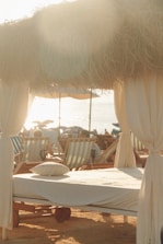 A serene beach scene with sunlight filtering through dried thatched roofing over a cabana set up. A neatly made sunbed with a pillow is in the foreground, surrounded by multiple beach chairs. The setting captures a relaxed, seaside atmosphere.