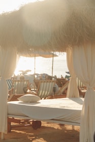 A serene beach scene with sunlight filtering through dried thatched roofing over a cabana set up. A neatly made sunbed with a pillow is in the foreground, surrounded by multiple beach chairs. The setting captures a relaxed, seaside atmosphere.