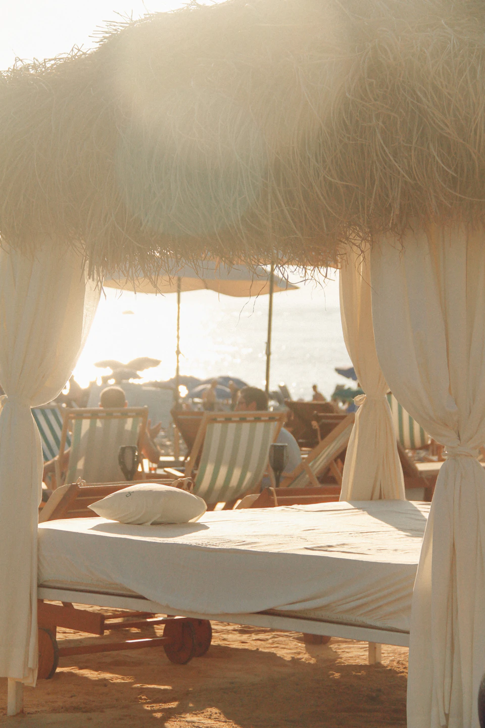 Beachgoers relax under a white canopy tent with lounge chairs and straw accents nearby