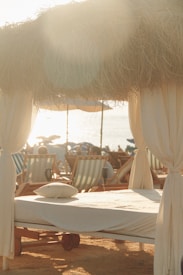 A serene beach scene with sunlight filtering through dried thatched roofing over a cabana set up. A neatly made sunbed with a pillow is in the foreground, surrounded by multiple beach chairs. The setting captures a relaxed, seaside atmosphere.