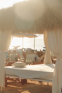 A serene beach scene with sunlight filtering through dried thatched roofing over a cabana set up. A neatly made sunbed with a pillow is in the foreground, surrounded by multiple beach chairs. The setting captures a relaxed, seaside atmosphere.