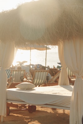 A serene beach scene with sunlight filtering through dried thatched roofing over a cabana set up. A neatly made sunbed with a pillow is in the foreground, surrounded by multiple beach chairs. The setting captures a relaxed, seaside atmosphere.