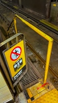 A subway train track area with a safety barrier and a warning sign. The yellow sign features a symbol indicating danger with the text 'Danger Do Not Enter' and an image of a train. The platform edge is marked with yellow tactile paving and a red strip labeled 'Edge.'