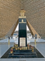A tall, dark monument stands in the center with inscriptions on its surface. The monument is surrounded by neatly arranged potted plants at its base. The structure is set within an architectural space featuring intricate geometric patterns on the walls and mirrored flooring that reflects light.