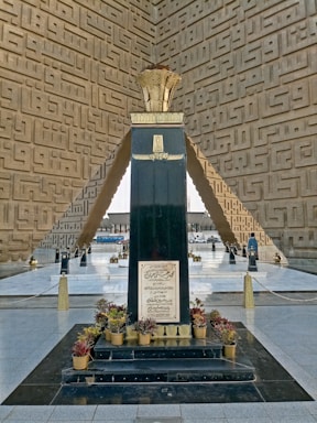 A tall, dark monument stands in the center with inscriptions on its surface. The monument is surrounded by neatly arranged potted plants at its base. The structure is set within an architectural space featuring intricate geometric patterns on the walls and mirrored flooring that reflects light.
