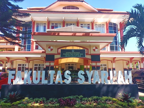 A large, multi-story building with an architectural style featuring prominent vertical and horizontal lines, large windows, and a pitched roof. In front of the building, there is a wide sign with large white letters spelling 'FAKULTAS SYARIAH'. The signage is situated on a black base with small plants in front. The entrance to the building has a sign above the door with text, and the building is flanked by tall palm trees.