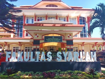 A large, multi-story building with an architectural style featuring prominent vertical and horizontal lines, large windows, and a pitched roof. In front of the building, there is a wide sign with large white letters spelling 'FAKULTAS SYARIAH'. The signage is situated on a black base with small plants in front. The entrance to the building has a sign above the door with text, and the building is flanked by tall palm trees.