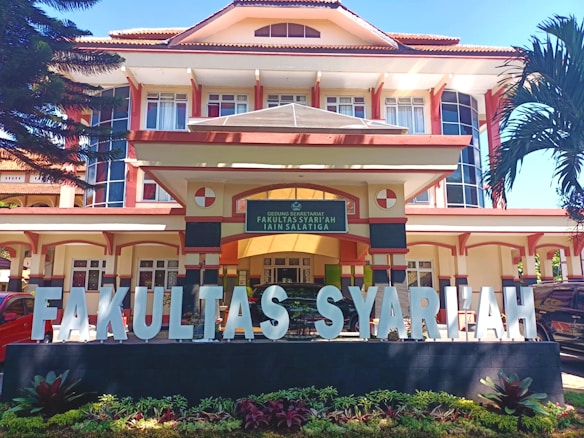 A large, multi-story building with an architectural style featuring prominent vertical and horizontal lines, large windows, and a pitched roof. In front of the building, there is a wide sign with large white letters spelling 'FAKULTAS SYARIAH'. The signage is situated on a black base with small plants in front. The entrance to the building has a sign above the door with text, and the building is flanked by tall palm trees.