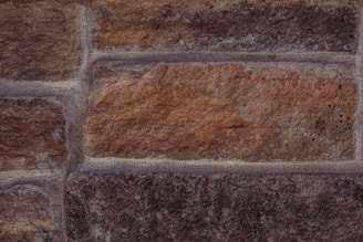A close-up view of a brick or stone wall with various shades of brown and gray. The wall consists of rectangular blocks with visible textures and mortar lines.