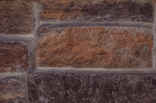A close-up view of a brick or stone wall with various shades of brown and gray. The wall consists of rectangular blocks with visible textures and mortar lines.