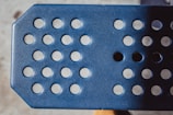 A close-up view of a blue perforated metal surface with circular holes arranged in rows. The lighting creates a play of shadows across the textured surface.