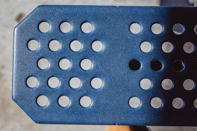 A close-up view of a blue perforated metal surface with circular holes arranged in rows. The lighting creates a play of shadows across the textured surface.