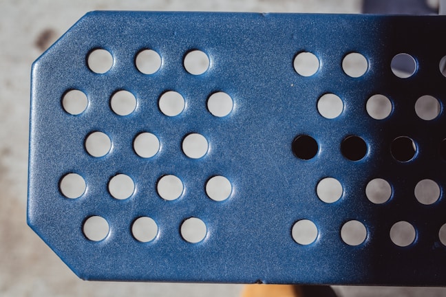 A close-up view of a blue perforated metal surface with circular holes arranged in rows. The lighting creates a play of shadows across the textured surface.