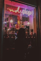 A dimly lit storefront window display features a large toy soldier surrounded by smaller toy soldiers. Neon lights in pink and purple cast a glow, with a sign overhead. The silhouette of a person looking into the window creates a mysterious vibe.