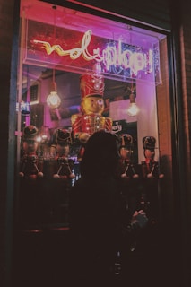 A dimly lit storefront window display features a large toy soldier surrounded by smaller toy soldiers. Neon lights in pink and purple cast a glow, with a sign overhead. The silhouette of a person looking into the window creates a mysterious vibe.
