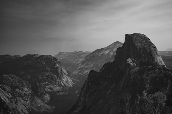 A dramatic black and white photograph of a mountainous landscape with rugged peaks and deep valleys. The prominent feature is a steep, expansive rock formation dominating the right side of the image. The sky is overcast, casting soft shadows over the terrain, enhancing the textures of the rocks.