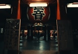 A large red lantern with bold white and black characters hangs in the entrance to a traditional temple area. The setting appears to be dimly lit, with the lantern providing a focal point. On either side, there are signs with Japanese text and people can be seen walking in the background.