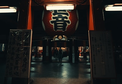 A large red lantern with bold white and black characters hangs in the entrance to a traditional temple area. The setting appears to be dimly lit, with the lantern providing a focal point. On either side, there are signs with Japanese text and people can be seen walking in the background.
