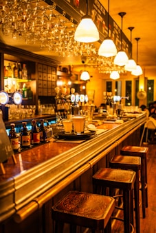 A warmly lit bar counter with several stools lined up along it. Glasses hang overhead, and a variety of bottles and taps are visible behind the counter. The atmosphere appears cozy and inviting, with soft, amber lighting casting a glow on the wooden surfaces.