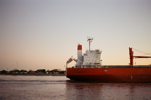 A large cargo ship with a red hull is sailing in calm waters near the coast. Trees and small buildings are visible in the background under a clear sky with a soft gradient from light blue to peach.