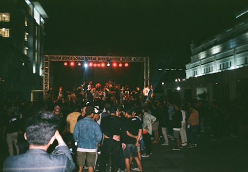 A large crowd of people is gathered at an outdoor event with a stage in the background. Red stage lights are illuminating a group of performers or speakers on stage. The scene is set at night, with buildings on either side visible under artificial lights.
