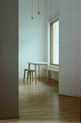 A minimalist room featuring a simple wooden table and stool set against a large window. The walls and floor are neutral in color, with exposed light bulbs hanging from the ceiling. The space appears open and uncluttered.